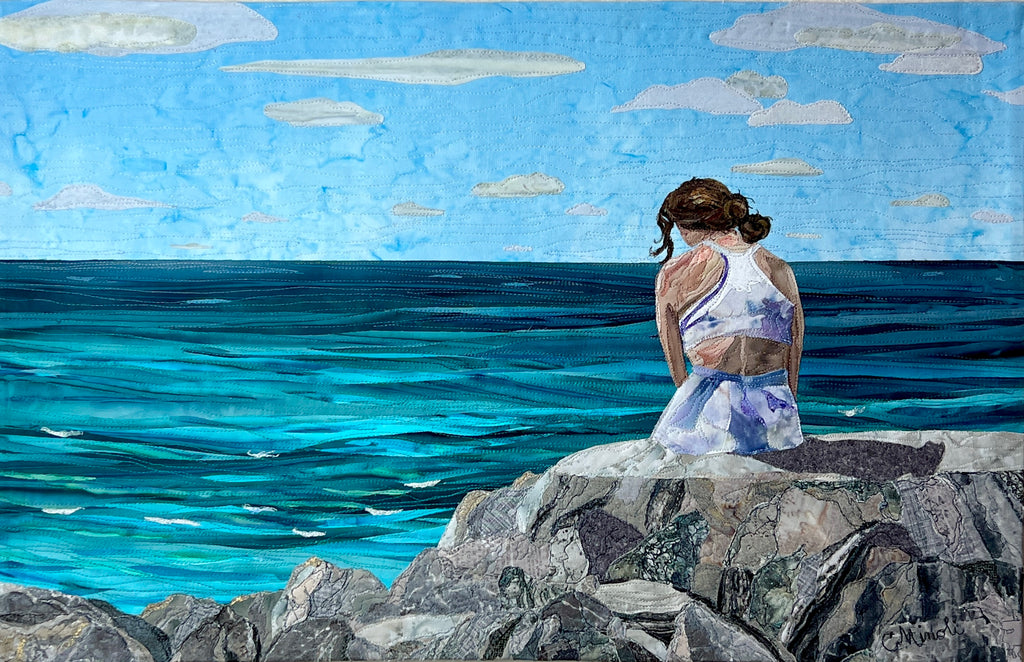 textile art of lady sitting on rocks by the ocean